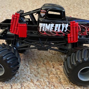 May include: A black monster truck toy with large black tires and red accents. The truck features the text "TIME FLYS" in white with red flames. The toy has a black chassis and a detailed design.