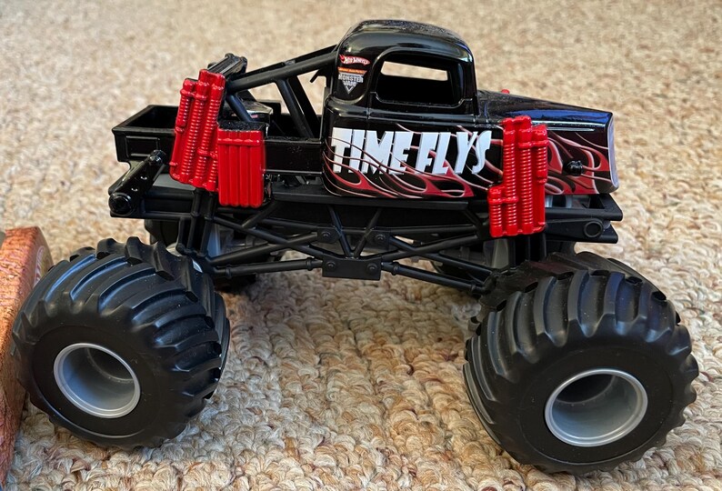 May include: A black monster truck toy with large black tires and red accents. The truck features the text "TIME FLYS" in white with red flames. The toy has a black chassis and a detailed design.