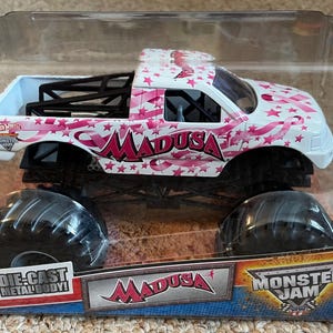 K&ouml;nnte beinhalten: Ein Monster Jam Truck aus Metallguss namens "Madusa" in einer transparenten Kunststoffverpackung. Der Truck ist wei&szlig; mit rosa Stern- und Bandgrafiken. Die Reifen sind schwarz.