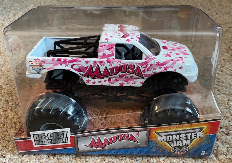 K&ouml;nnte beinhalten: Ein Monster Jam Truck aus Metallguss namens "Madusa" in einer transparenten Kunststoffverpackung. Der Truck ist wei&szlig; mit rosa Stern- und Bandgrafiken. Die Reifen sind schwarz.
