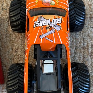 May include: Orange monster truck toy with black wheels. The truck features the text "BAD NEWS!" and a cartoon character with crossed newspapers. The words "YOU LOSE...TO" are also visible. The truck has a black chassis and a silver engine.