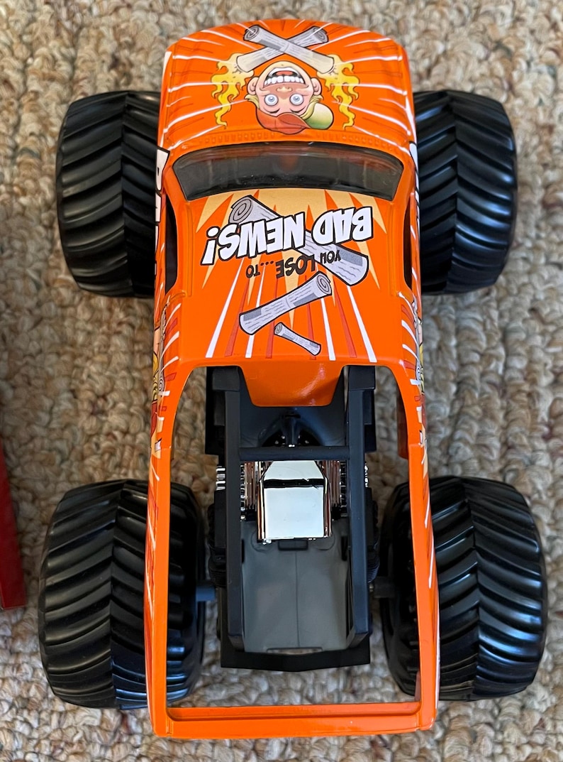 May include: Orange monster truck toy with black wheels. The truck features the text "BAD NEWS!" and a cartoon character with crossed newspapers. The words "YOU LOSE...TO" are also visible. The truck has a black chassis and a silver engine.