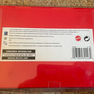 May include: A red cardboard box with a clear plastic cover. The box contains text including the Mattel logo, consumer information, and a barcode. The box is likely for a toy or collectible item.