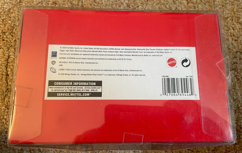 May include: A red cardboard box with a clear plastic cover. The box contains text including the Mattel logo, consumer information, and a barcode. The box is likely for a toy or collectible item.