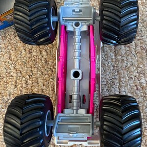 May include: A toy monster truck with large black tires and a gray and pink chassis. The tires have a deep tread pattern. The truck's body is gray and pink, with a detailed engine compartment.