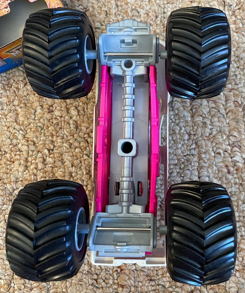 May include: A toy monster truck with large black tires and a gray and pink chassis. The tires have a deep tread pattern. The truck's body is gray and pink, with a detailed engine compartment.