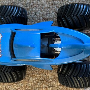 May include: A blue toy monster truck with black tyres. The truck is shaped like a shark and has the words "Shark Wreak" in red on the side. The tyres are large and have a deep tread.