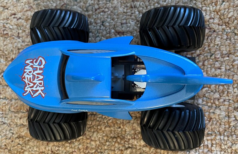 May include: A blue toy monster truck with black tyres. The truck is shaped like a shark and has the words "Shark Wreak" in red on the side. The tyres are large and have a deep tread.