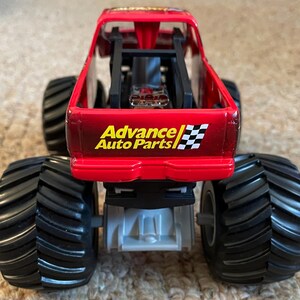 May include: A red toy monster truck with large black tires. The truck has the "Advance Auto Parts" logo and a checkered flag design on the back. The truck is a toy vehicle.