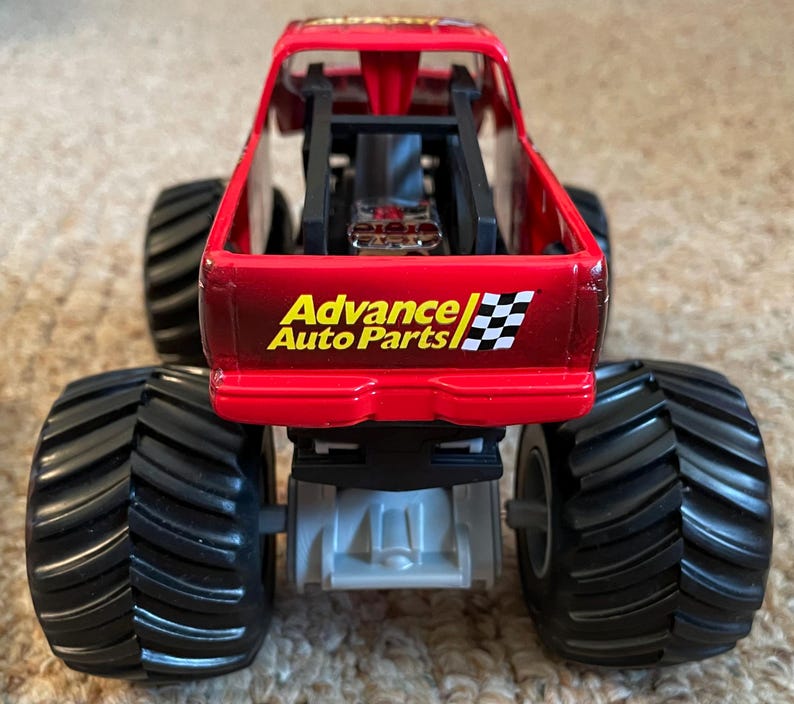 May include: A red toy monster truck with large black tires. The truck has the "Advance Auto Parts" logo and a checkered flag design on the back. The truck is a toy vehicle.