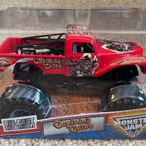 May include: A red die-cast metal body monster truck toy, "Captain's Curse", in a clear plastic package. The truck has large black tires and a black roll cage. The package features the Monster Jam logo.