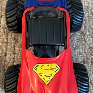 Op de afbeelding: Een rood, blauw en zwart Superman monstertruck speelgoed. De truck heeft grote zwarte banden en het Superman-logo op de motorkap en het dak. Het speelgoed is ontworpen met een rode cape-achtige voorkant.