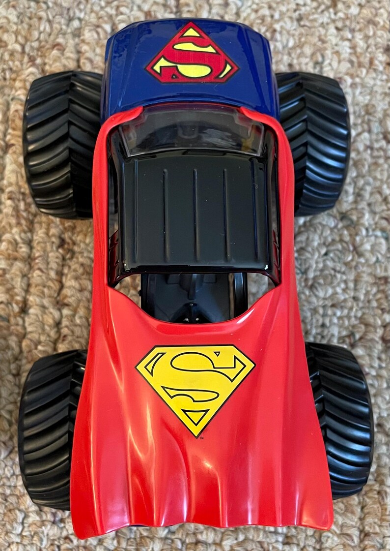 Op de afbeelding: Een rood, blauw en zwart Superman monstertruck speelgoed. De truck heeft grote zwarte banden en het Superman-logo op de motorkap en het dak. Het speelgoed is ontworpen met een rode cape-achtige voorkant.