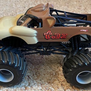 May include: A brown and tan monster truck toy with the word "taz" in red on the side. The truck has large black tires and a cartoon character design on the front.