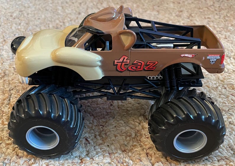 May include: A brown and tan monster truck toy with the word "taz" in red on the side. The truck has large black tires and a cartoon character design on the front.