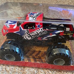 May include: A red, white, and blue Nitro Circus monster truck toy in a clear plastic package. The truck has large black tyres and the Nitro Circus logo. The packaging is red and the truck is in a clear plastic case.
