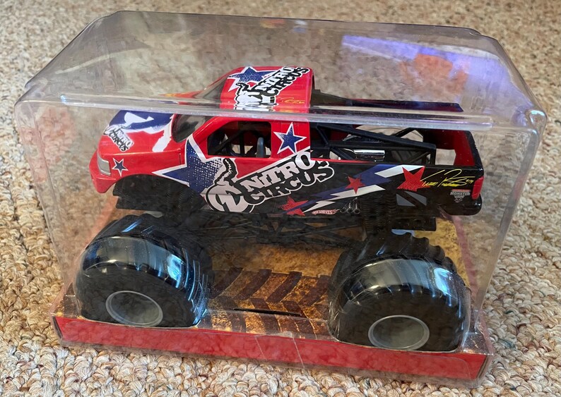May include: A red, white, and blue Nitro Circus monster truck toy in a clear plastic package. The truck has large black tyres and the Nitro Circus logo. The packaging is red and the truck is in a clear plastic case.