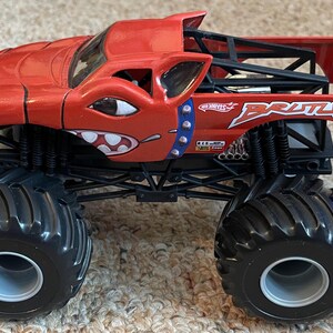 May include: A red Hot Wheels monster truck toy with black wheels and suspension. The truck has a cartoon face design and the name "Brutus". The truck has a roll cage and a bed.
