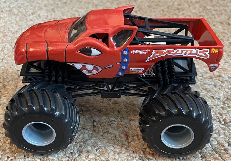 May include: A red Hot Wheels monster truck toy with black wheels and suspension. The truck has a cartoon face design and the name "Brutus". The truck has a roll cage and a bed.