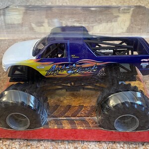 May include: A monster truck toy in a clear plastic package. The truck is primarily blue with white and yellow accents. The text "Aftershock" is visible on the side. The truck has large black tires and is on a red and brown base.