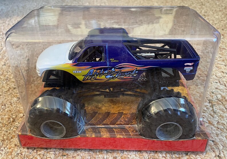 May include: A monster truck toy in a clear plastic package. The truck is primarily blue with white and yellow accents. The text "Aftershock" is visible on the side. The truck has large black tires and is on a red and brown base.