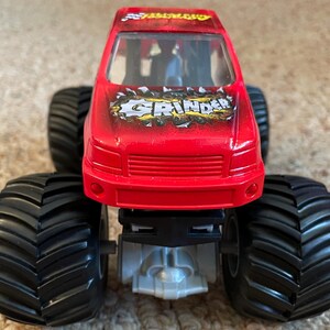 May include: A red toy monster truck with large black tires. The truck has the word "GRINDER" in a stylized font on the hood. The truck is a toy vehicle, likely for children.