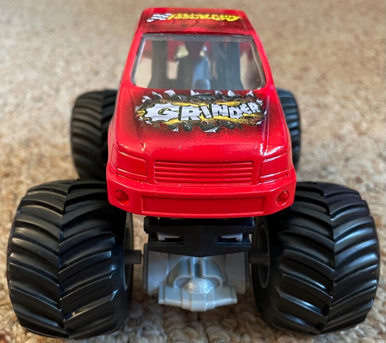 May include: A red toy monster truck with large black tires. The truck has the word "GRINDER" in a stylized font on the hood. The truck is a toy vehicle, likely for children.