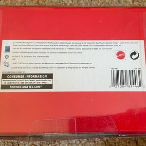 Może przedstawiać: Czerwone kartonowe pudełko z przezroczystym plastikowym opakowaniem. Pudełko ma biały tekst i kod kreskowy. Tekst zawiera "Consumer Information" i "Service.Mattel.com". Pudełko leży na beżowym dywanie.