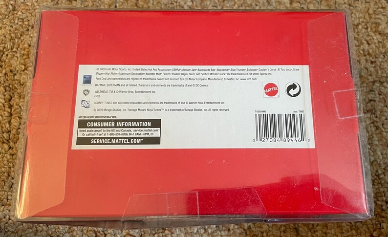 Może przedstawiać: Czerwone kartonowe pudełko z przezroczystym plastikowym opakowaniem. Pudełko ma biały tekst i kod kreskowy. Tekst zawiera "Consumer Information" i "Service.Mattel.com". Pudełko leży na beżowym dywanie.