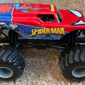 May include: A red and blue Spider-Man monster truck toy with large black tires. The truck features the Spider-Man logo and graphics, with the words "SPIDER-MAN" and "MONSTER JAM" visible.