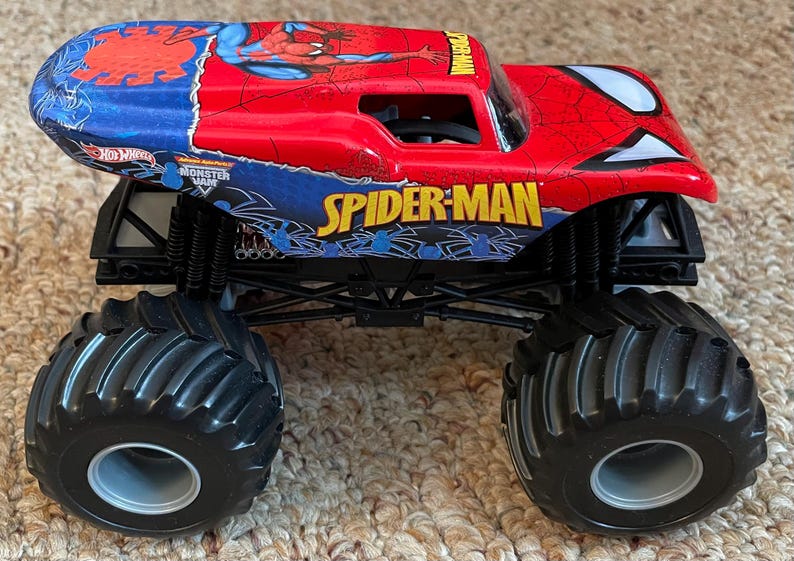 May include: A red and blue Spider-Man monster truck toy with large black tires. The truck features the Spider-Man logo and graphics, with the words "SPIDER-MAN" and "MONSTER JAM" visible.