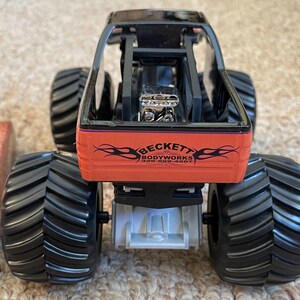 May include: A toy monster truck with large black tires and a black frame. The truck has an orange body with the text "BECKETT BODYWORKS" and a flame design. The engine is visible. The toy is on a light brown surface.