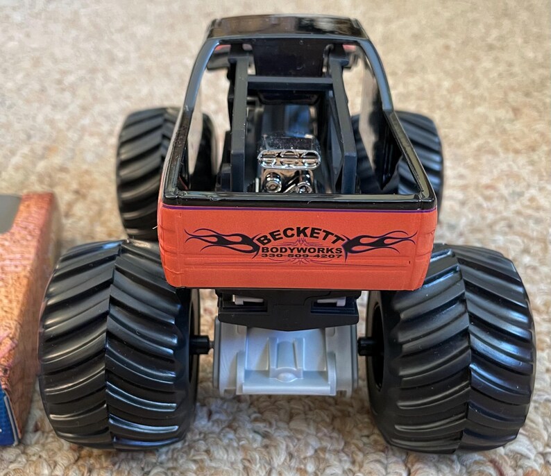 May include: A toy monster truck with large black tires and a black frame. The truck has an orange body with the text "BECKETT BODYWORKS" and a flame design. The engine is visible. The toy is on a light brown surface.