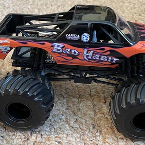 May include: A black and orange toy monster truck with the words "Bad Habit" and flame graphics. The truck has large black tires and a black roll cage. The Hot Wheels logo is visible.