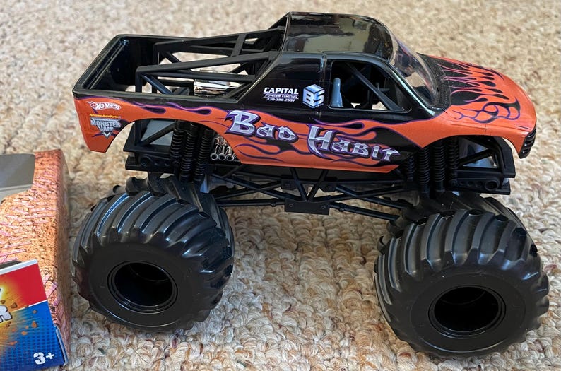 May include: A black and orange toy monster truck with the words "Bad Habit" and flame graphics. The truck has large black tires and a black roll cage. The Hot Wheels logo is visible.