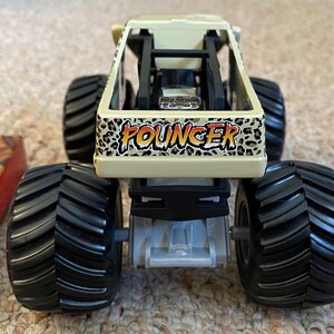 May include: A toy monster truck with large black tires and a tan body. The truck features the word "FOUNGER" in a leopard print design on the front. The truck is designed for off-road play.