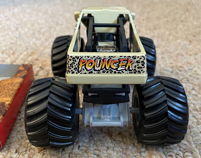 May include: A toy monster truck with large black tires and a tan body. The truck features the word "FOUNGER" in a leopard print design on the front. The truck is designed for off-road play.