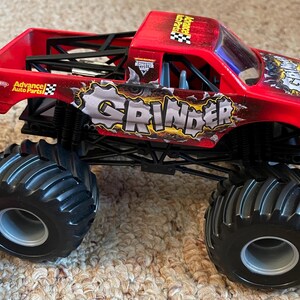May include: A red toy monster truck with the word "GRINDER" in large letters on the side. The truck has large black tires and the words "Advance Auto Parts" are visible. The truck is a toy.