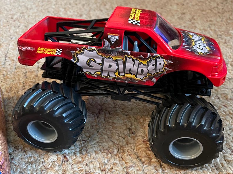 May include: A red toy monster truck with the word "GRINDER" in large letters on the side. The truck has large black tires and the words "Advance Auto Parts" are visible. The truck is a toy.