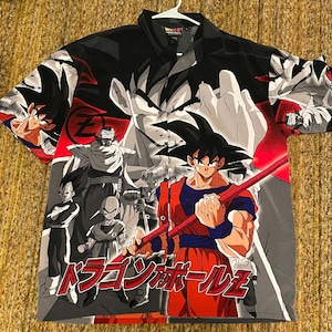 May include: Button-up shirt featuring Dragon Ball Z characters in a vibrant design. The shirt showcases Goku in his signature orange and blue attire, with Japanese text and other characters in a dynamic composition. The shirt is on a white hanger.