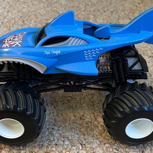 May include: A blue monster truck toy with a shark design. The truck has large black tyres with white rims, a shark fin, and the text "Capt. Carlyle". The shark's mouth is open, revealing white teeth.