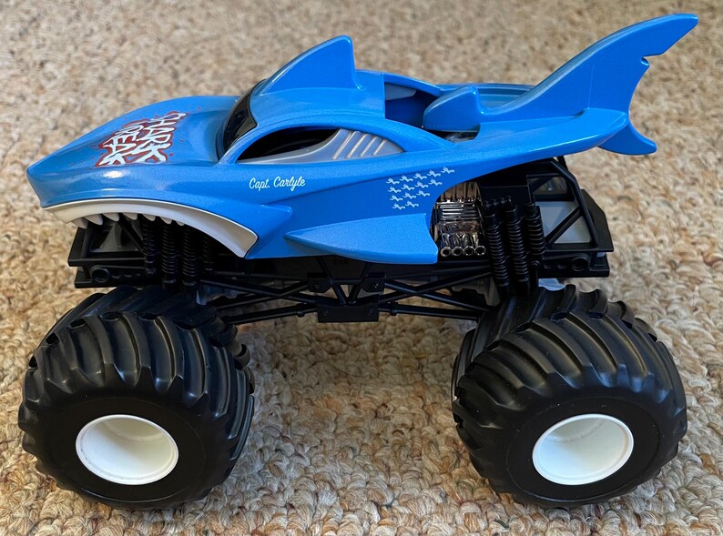May include: A blue monster truck toy with a shark design. The truck has large black tyres with white rims, a shark fin, and the text "Capt. Carlyle". The shark's mouth is open, revealing white teeth.