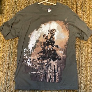 May include: Olive green t-shirt featuring a Marvel comic book illustration. The graphic depicts Spider-Man, Wolverine, and another character on a rooftop with a cityscape backdrop. The Marvel logo is visible on the shirt's collar.