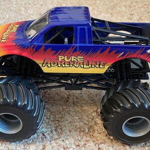 Może przedstawiać: Niebiesko-ż&oacute;łto-czerwona zabawka monster truck z napisem "Pure Adrenaline". Ciężar&oacute;wka ma duże czarne opony i logo Hot Wheels Monster Jam.