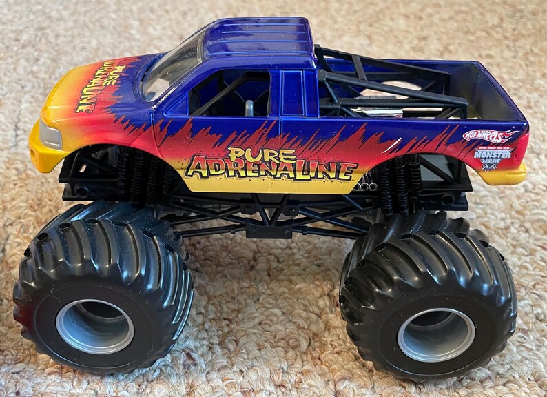 Może przedstawiać: Niebiesko-ż&oacute;łto-czerwona zabawka monster truck z napisem "Pure Adrenaline". Ciężar&oacute;wka ma duże czarne opony i logo Hot Wheels Monster Jam.