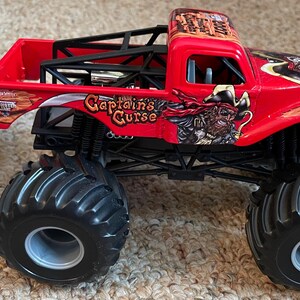 May include: A red toy monster truck with black tires and a black roll cage. The truck has the words "Captain's Curse" and a pirate graphic on the side. The truck is sitting on a carpeted surface.