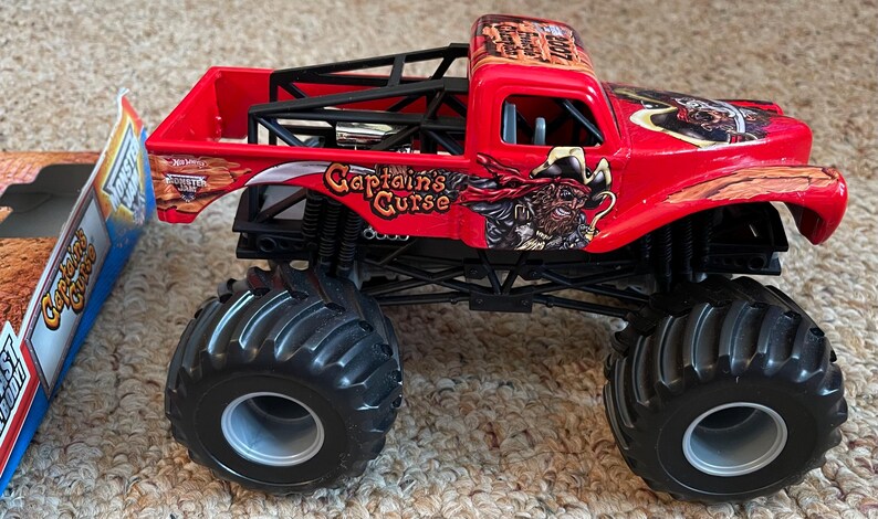 May include: A red toy monster truck with black tires and a black roll cage. The truck has the words "Captain's Curse" and a pirate graphic on the side. The truck is sitting on a carpeted surface.