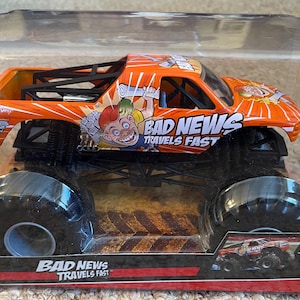 May include: Orange monster truck toy with black wheels and a black roll cage. The truck has the words "BAD NEWS TRAVELS FAST" and a cartoon graphic. The toy is in a clear plastic package.