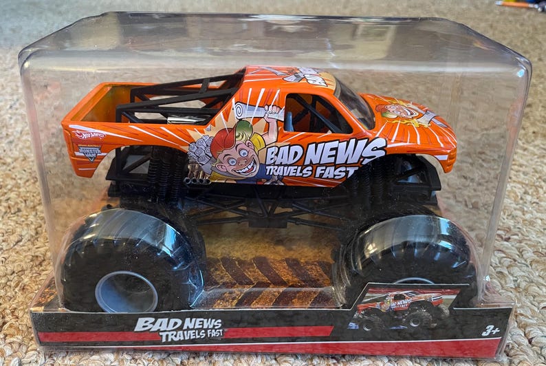 May include: Orange monster truck toy with black wheels and a black roll cage. The truck has the words "BAD NEWS TRAVELS FAST" and a cartoon graphic. The toy is in a clear plastic package.