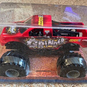 May include: A red and black toy monster truck named "Grinder" in a clear plastic package. The truck has large black tires and the words "Advance Auto Parts" are visible. The truck is a toy vehicle.